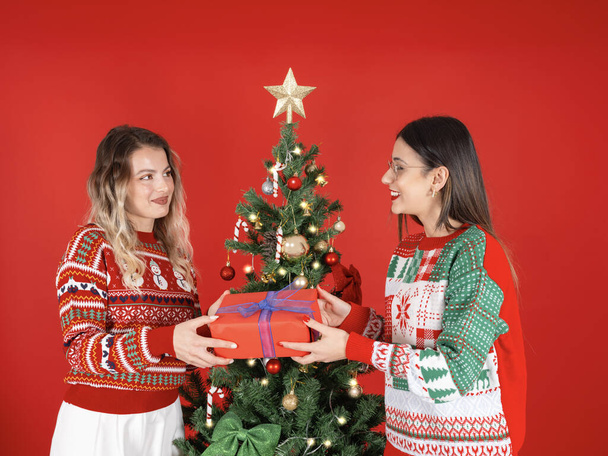 Giving Geschenk-Box, Porträt von zwei jungen kaukasischen Mädchen besten Freundinnen Schwestern geben Geschenk-Box. In der Nähe des geschmückten Weihnachtsbaums stehen und im kuscheligen Pullover ein frohes neues Jahr begrüßen. Roter Hintergrund. - Foto, Bild