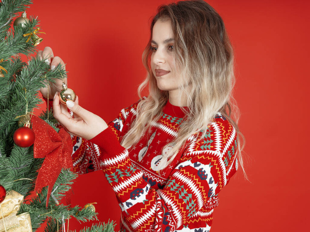 クリスマスツリーを飾る,サイドビュークリスマスツリーを飾る若い美しい女性の画像を閉じます. 着心地の良い暖かいセーターを着用してください. 赤いスタジオの背景の上に立ってください. ハッピーニューイヤーアクティビティコンセプト. - 写真・画像