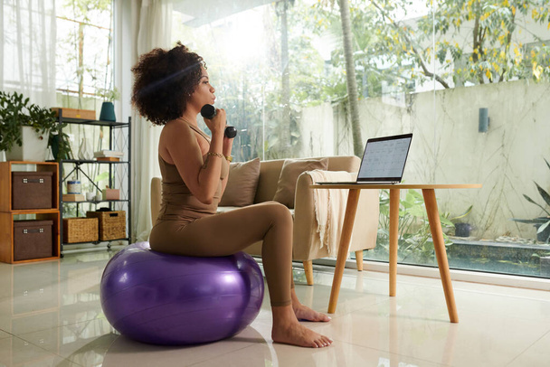Frau trainiert mit Kurzhanteln auf Fitnessball, wenn sie ihren Kalender auf dem Laptop checkt - Foto, Bild