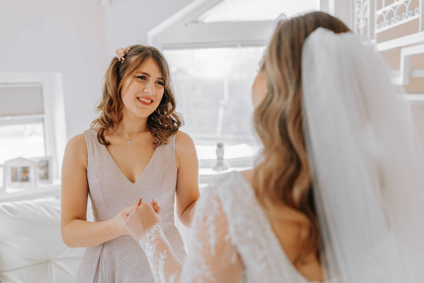A friend helps the bride fasten her dress. A woman helps her friend fasten the buttons on the back of her wedding dress. The morning of the bride, the creation of a family, an important event. - Foto, Imagen