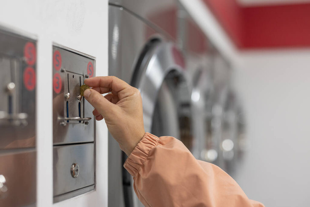 Mãos de uma pessoa vista inserindo uma moeda ou token na lavanderia. .. Máquinas de lavar ao fundo, mão feminina em primeiro plano. - Foto, Imagem