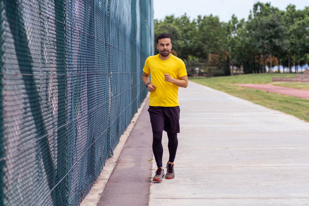Człowiek z brodą w wygodnym dresie biegnie wzdłuż drogi, mijając wysoki płot siatkowy na boisku sportowym w parku miejskim, angażując się w działania na rzecz zdrowia i rozwoju ciała.  - Zdjęcie, obraz