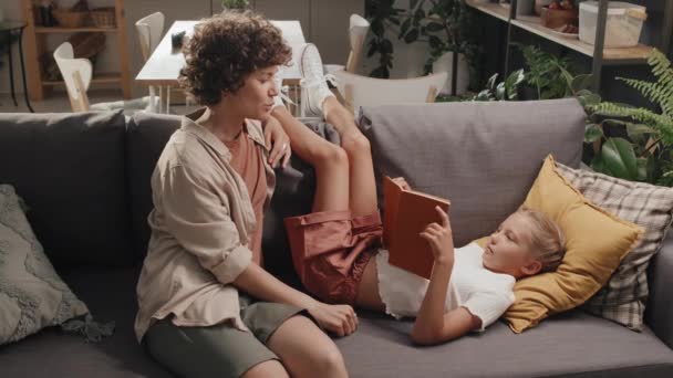 Αργή κίνηση πλάνο της σύγχρονης μητέρας και της κόρης ξοδεύουν χρόνο μαζί στο σπίτι χαλαρώνοντας στον καναπέ στο σαλόνι και συζητώντας το βιβλίο - Πλάνα, βίντεο