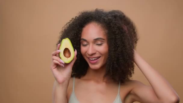 Smiling African American model beautiful woman girl with gorgeous hair shake head curls enjoy eco shampoo cosmetic conditioner curly hairstyle in beige ad advertise background holding half avocado - Footage, Video