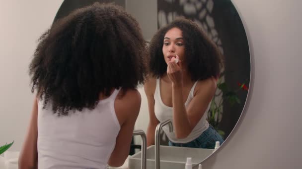 Genç Afrikalı Amerikalı kadın sabah makyajı hazırlıyor aynanın yansımasına bakıyor nemlendirici koruyucu dudak kremi ruj sürüyor organik doğal ürün. - Video, Çekim