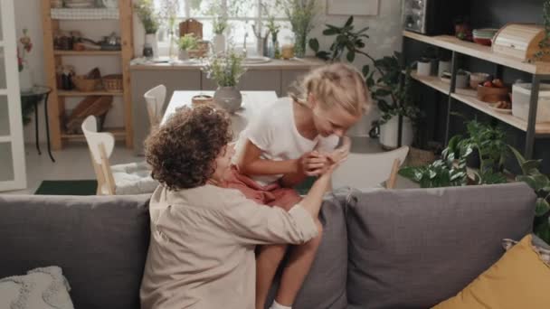 Zeitlupenaufnahme einer verspielten Mutter, die auf der Couch sitzt und Spaß daran hat, ihre Tochter zu kitzeln - Filmmaterial, Video