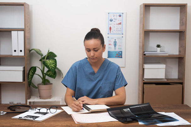 Tıbbi İnceleme: Ofis masasında, mavi üniformalı doktor tıbbi belgelerin ve test sonuçlarının eksiksiz bir şekilde incelenmesine odaklanarak,.  - Fotoğraf, Görsel