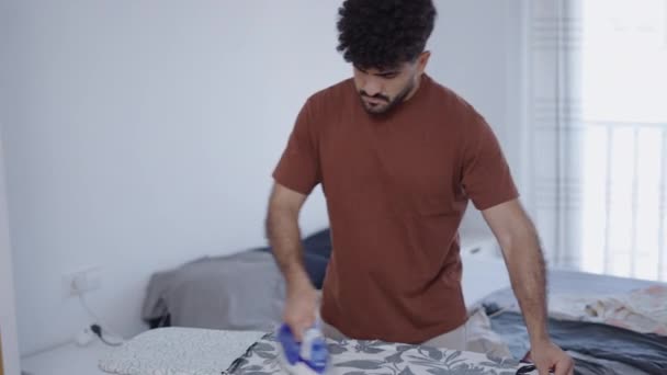 chico latino planchando su camisa impresa en su habitación - 4K Horizontal video - Imágenes, Vídeo