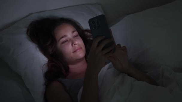 Een late avond scène: een vrouw, gebonden aan bed, gefascineerd door haar telefoon scherm, belichaamt de uitdagingen van slapeloosheid en smartphone afhankelijkheid.  - Video