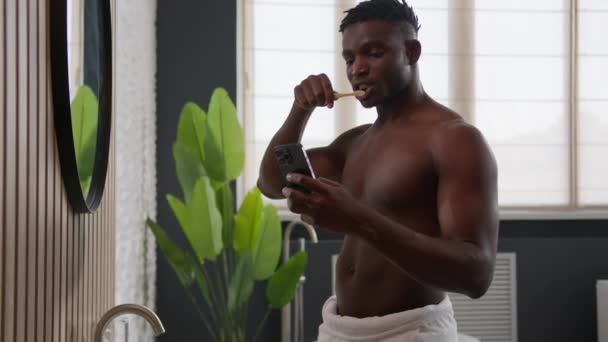 Afrikkalainen amerikkalainen mies harjaus hampaat ja vieritys matkapuhelin kotona kylpyhuone kiireinen ajanhallinta etninen mies suuhygienia aamu järkyttynyt kaveri lukea uskomattomia uutisia älypuhelin tuntuu shokki ihme - Materiaali, video