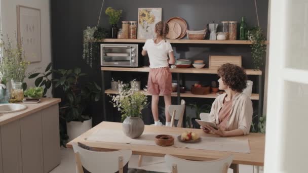 Moderni esiteini tyttö tekee askareita keittiössä pyyhkimällä hyllyjä, kun hänen äitinsä työskentelee digitaalisella tabletilla - Materiaali, video