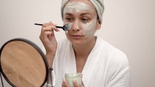 自宅のスパ:バスローブでは,彼女は彼女の有機顔の治療を楽しんで,鏡を見つめ,粘土マスクを適用するためにブラシを使用しています.  - 映像、動画