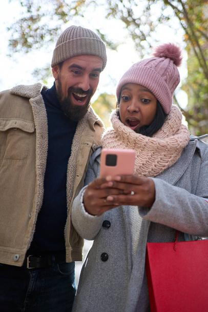 Vídeo vertical. pareja joven interracial en el amor usando el teléfono móvil con la cara sorprendida disfrutando de la temporada de vacaciones de invierno en las calles de la ciudad y sosteniendo bolsas de regalo mientras se celebra la Navidad al aire libre - Foto, Imagen