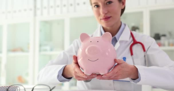Woman doctor with stethoscope shows pink piggy bank with money savings inside sitting at desk in hospital. Concept of charitable foundation to support sick people - Footage, Video