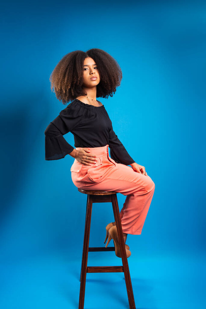 Piękna młoda kobieta z czarnymi włosami siedząca na drewnianym stołku. Izolacja na niebieskim tle. - Zdjęcie, obraz
