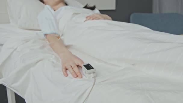 Messa a fuoco selettiva panning shot di paziente donna caucasica sdraiata priva di sensi in letto d'ospedale che riceve fluidi attraverso la linea endovenosa e indossa pulsossimetro sulla punta delle dita - Filmati, video