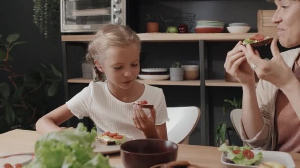 Średnie ujęcie nowoczesnej mamy i córki siedzących przy stole w kuchni cieszących się razem śniadaniem - Materiał filmowy, wideo
