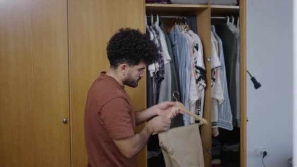 Homem latino abre seu guarda-roupa e tira um par de calças de camelo e uma camisa de pólo branco - vídeo 4K Horizontal - Filmagem, Vídeo
