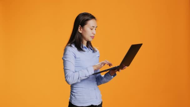 Casual person navigating online on laptop in studio, looking for inspiration on wireless pc against orange background. Asian woman browsing webpages on internet, search project. - Footage, Video