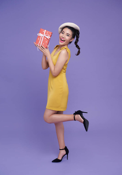 Ευτυχισμένο χαμόγελο διασκέδαση ασιατικό κορίτσι σε κίτρινο φόρεμα κρατώντας το παρόν κουτί αυξήσει το πόδι απομονώνονται σε μωβ φόντο. - Φωτογραφία, εικόνα