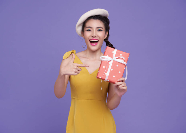 felice sorridente divertimento ragazza asiatica in abito giallo puntando il dito per presentare la scatola isolata su sfondo viola. - Foto, immagini