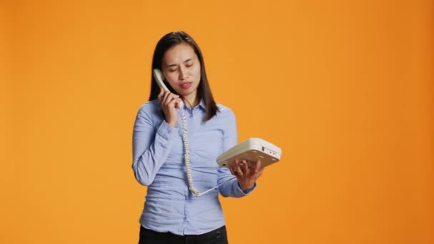 Persona asiática haciendo una llamada remota en el teléfono fijo mientras habla con alguien en la antigua línea. Mujer segura manejando el teléfono de oficina con cable, interactuando en el estudio de forma remota. - Imágenes, Vídeo