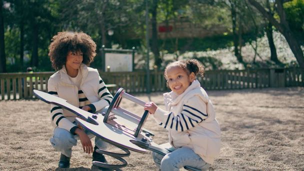 Meisje springt op schommel op zonnige openbare speelplaats. Speels Afrikaans Amerikaans kind geniet van spelen op schommelschommel buiten. Gelukkig lachende moeder kijken op zorgeloze schattige dochter. Vreugde concept - Foto, afbeelding