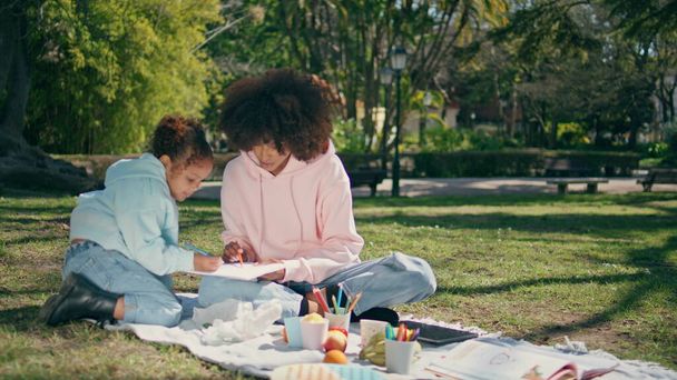 Kreative Familie beim Picknick auf schönem grünen Rasen am sonnigen Wochenende. Entspannte afrikanisch-amerikanische Mutter beim Malen auf Papier mit ihrer niedlichen kleinen Tochter, die Spaß an der Kunst hat. Fröhliches Mutti-Kind schafft Bild. - Foto, Bild