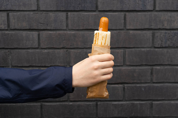 Ręczne trzymanie hot-doga na Black Street Wall Tło. Kiełbasa w opakowaniu papierowym, Street Fast Food z najbliższego sklepu, Hotdog na czarnym tle - Zdjęcie, obraz