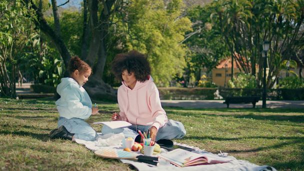 Family sitting picnic blanket on sunny green nature having fun together. Cute african american child drawing on paper with happy curly mother. Relaxed little girl enjoying creativity on meadow grass. - Photo, Image