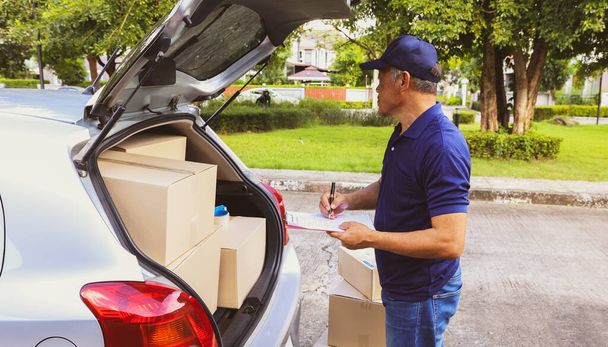Home delivery business : Senior male employee stands at the back of the truck, recording the customer's parcel delivery list and cardboard box products on a clipboard for accurate delivery. - Photo, Image
