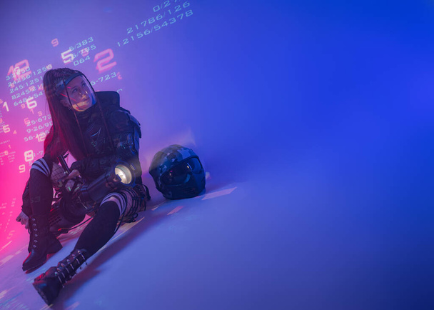 Kobieta w futurystycznym czarnym garniturze taktycznym leży na podłodze, w kasku motocyklowym i ogromnym projektorze leżącym obok niej, na tle projektowanych symboli cyfrowych. - Zdjęcie, obraz