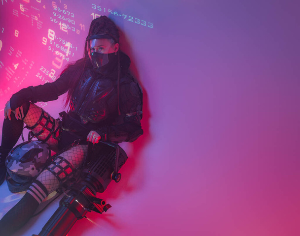 Nainen futuristisessa taktisessa mustassa puvussa makaa lattialla massiivinen projektori vierellään, taustalla ennustetut digitaaliset symbolit, kopiotilaa - Valokuva, kuva
