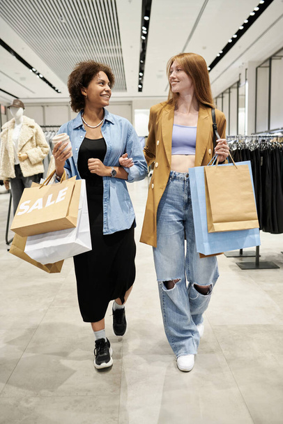 Tiro vertical completo de dos amigos sonrientes saliendo de la tienda llevando bolsas con compras - Foto, imagen