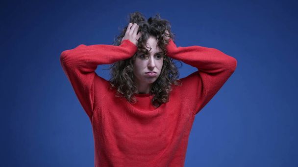 赤いセーターで青い背景に不安と絶望で彼女の髪を引っ張っている不思議な若い女性. 気になる20代女性 - 写真・画像