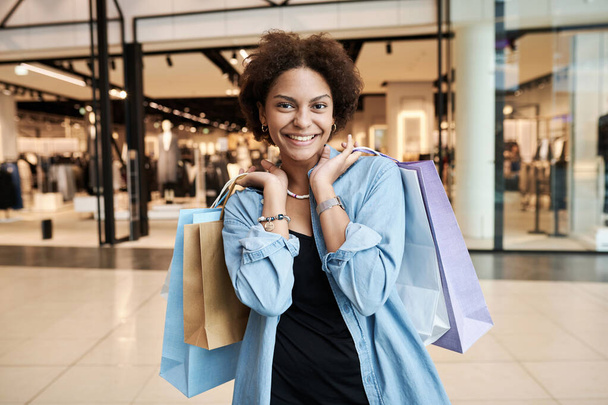 Feliz joven afroamericana chica posando con bolsas de papel con tienda de ropa en el fondo - Foto, imagen