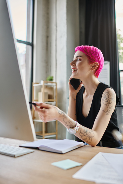 Neşeli dövmeli, sıradan giyinmiş iş kadını bilgisayarla uğraşırken telefonla konuşuyor. - Fotoğraf, Görsel