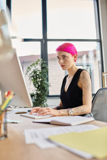 mujer de pelo rosa enfocada con tatuajes mirando su computadora mientras trabaja duro, negocio - Foto, imagen