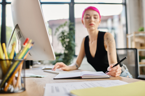 foto borrosa de la mujer de negocios de pelo rosa tomando notas mientras trabaja duro, se centran en su computadora - Foto, Imagen