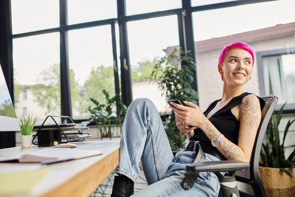 alegre joven mujer de negocios con el pelo rosa y tatuajes sentado y sonriendo en su escritorio en la oficina - Foto, Imagen