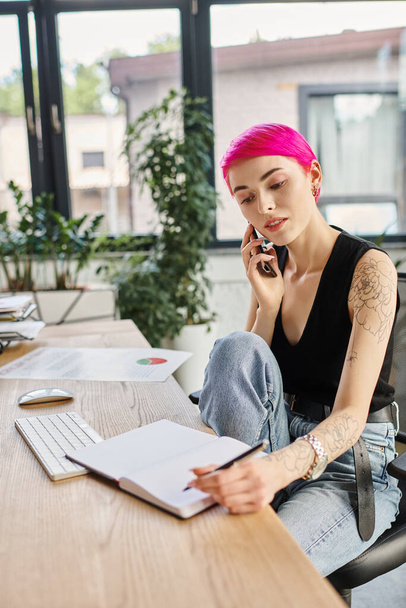 junge tätowierte Geschäftsfrau mit rosa Haaren in urbaner Kleidung macht sich Notizen und telefoniert - Foto, Bild