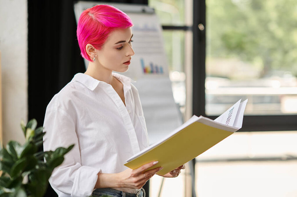 στοχαστική νεαρή επιχειρηματίας με ροζ κοντά μαλλιά σε casual ενδυμασία κοιτάζοντας τις σημειώσεις της στο γραφείο - Φωτογραφία, εικόνα