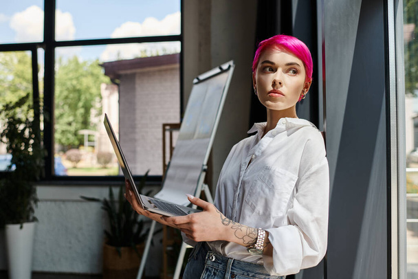 mulher de cabelos rosa pensativo com tatuagens segurando laptop e olhando para o escritório, conceito de negócio - Foto, Imagem