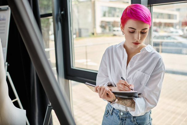 vrij pensive roze harige vrouw in wit shirt het maken van notities terwijl op kantoor, business concept - Foto, afbeelding