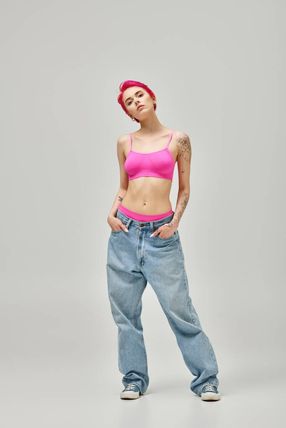 mujer atractiva con el pelo corto de color rosa y tatuajes en la parte superior de la cosecha de color rosa y jeans con las manos en los bolsillos - Foto, Imagen