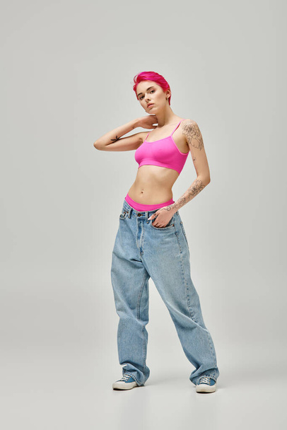 atraktivní mladá žena s růžovými vlasy a tetování ve stylové plodiny top pózování na šedém pozadí - Fotografie, Obrázek
