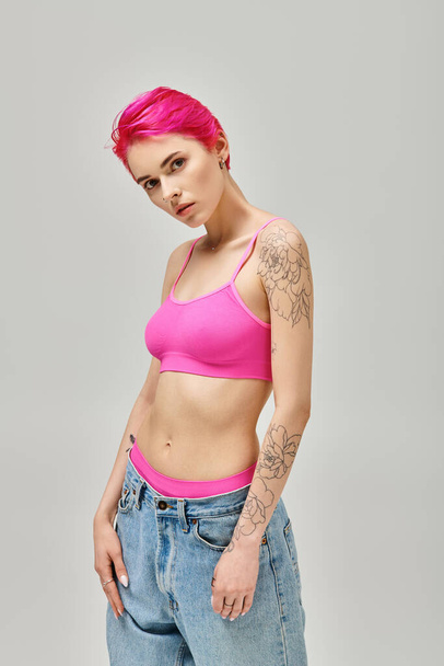 atraktivní krátkovlasá žena s tetováním v módní růžové střeše a džíny pózující na šedém pozadí - Fotografie, Obrázek