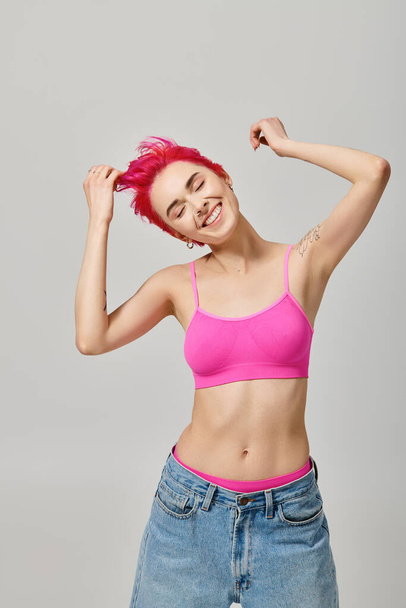 heureuse femme aux cheveux roses avec des tatouages posant avec les yeux fermés sur fond gris, concept de mode - Photo, image