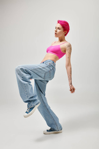 donna dai capelli rosa in elegante crop top e jeans in posa in movimento su sfondo grigio, concetto di moda - Foto, immagini