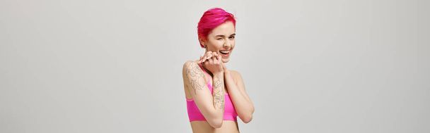 podekscytowana młoda kobieta z różowymi krótkimi włosami pozuje w górę i mruga na szarym tle, baner - Zdjęcie, obraz